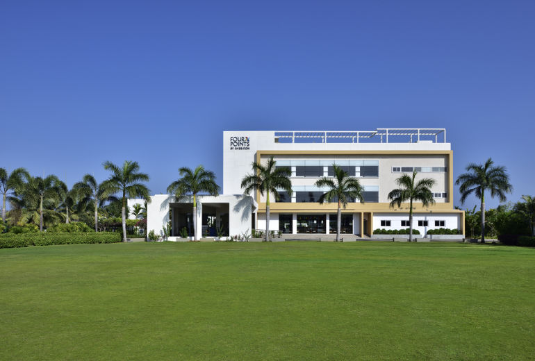A modern hotel building with a flat roof, labeled "Four Points by Sheraton," surrounded by palm trees. The foreground features a large, well-maintained grassy lawn under a clear blue sky.