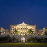 a large white building with a lawn and a walkway with Falaknuma Palace in the background