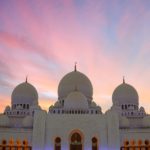 a large white building with domes and a pink and blue sky with Sheikh Zayed Mosque in the background
