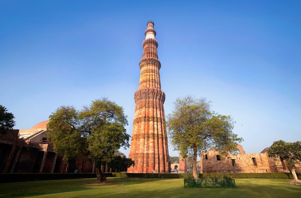 a tall brick tower with trees and grass with Qutub Minar in the background