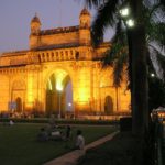 a large stone arch with people sitting on grass with Gateway of India in the background