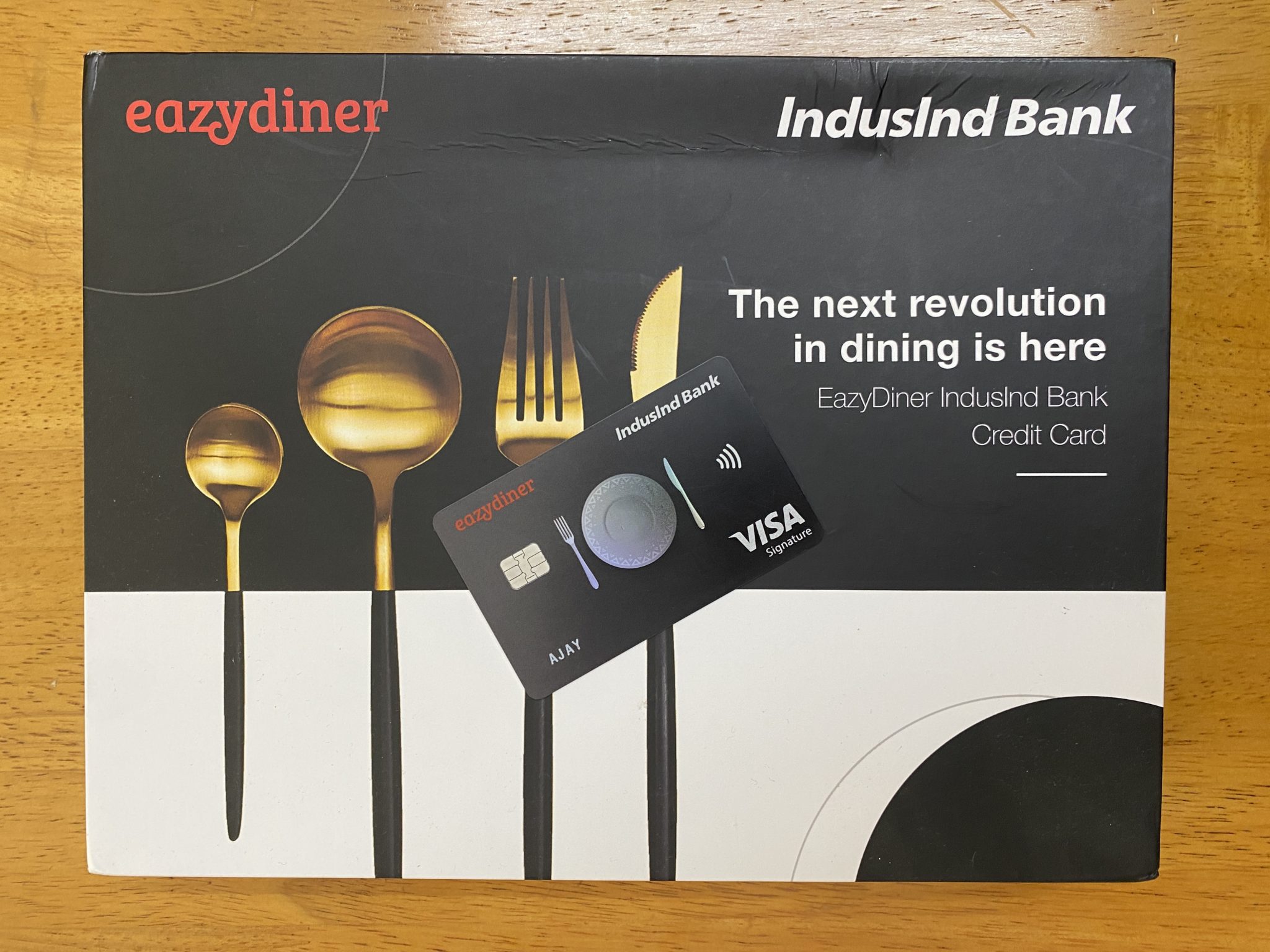 The image shows a promotional package for the EazyDiner IndusInd Bank Credit Card. The background features a design with golden cutlery, including spoons and a fork. The text on the package reads, "The next revolution in dining is here. EazyDiner IndusInd Bank Credit Card." A physical credit card is placed on top of the package, displaying the EazyDiner and IndusInd Bank logos, along with the Visa Signature logo.