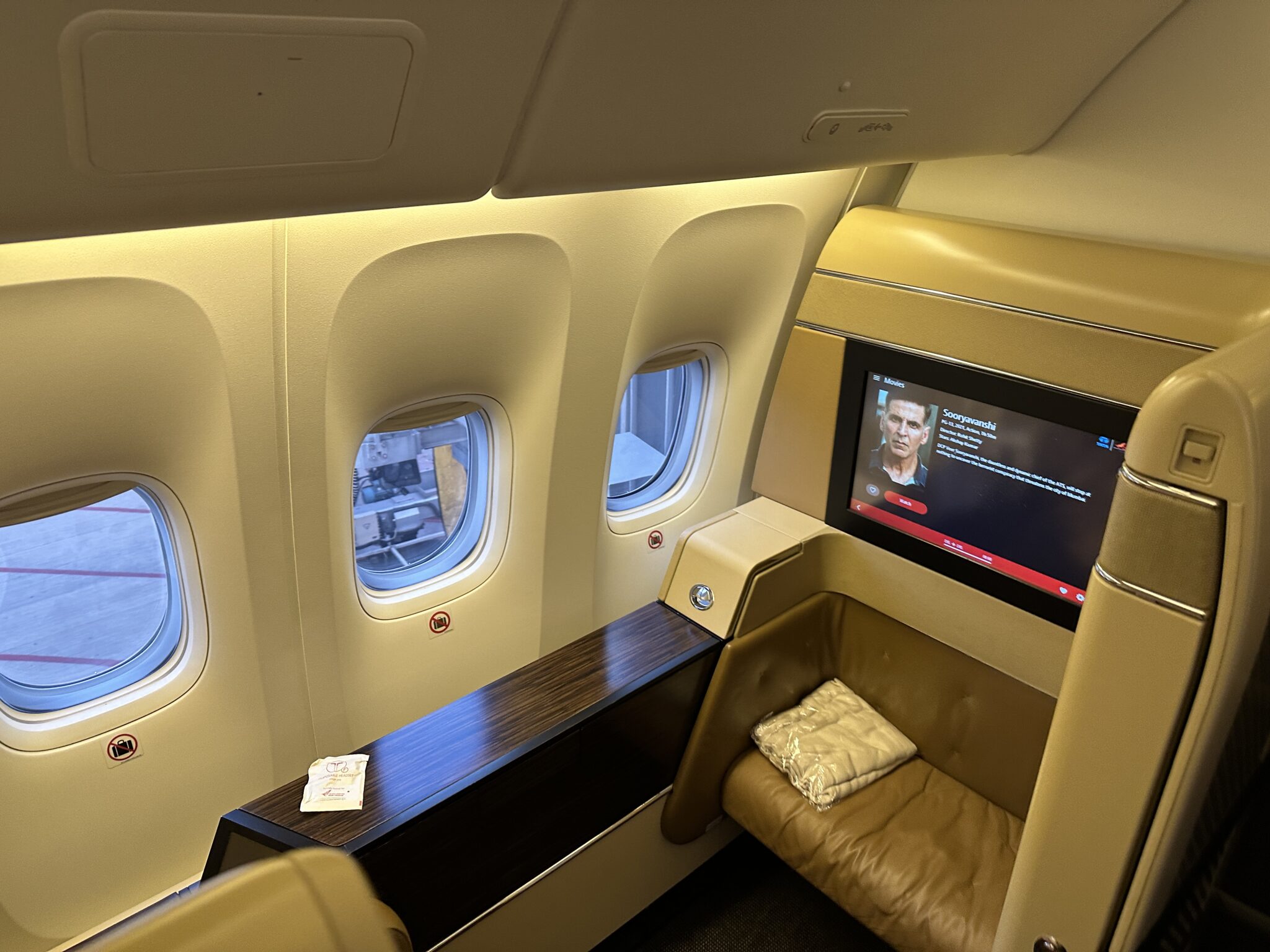 This image shows a luxurious first-class airplane cabin with a large, comfortable seat and a personal entertainment screen displaying a movie selection. There are three windows along the wall, and a small table with a packaged blanket or pillow on the seat. The cabin has a sleek, modern design with beige and brown tones.