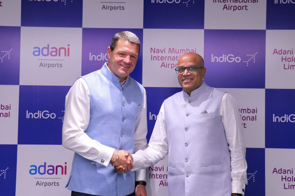 Two men are shaking hands and smiling at the camera. They are both wearing light blue vests over white shirts. The background features a checkered pattern with logos and text, including "adani Airports," "IndiGo," and "Navi Mumbai International Airport."