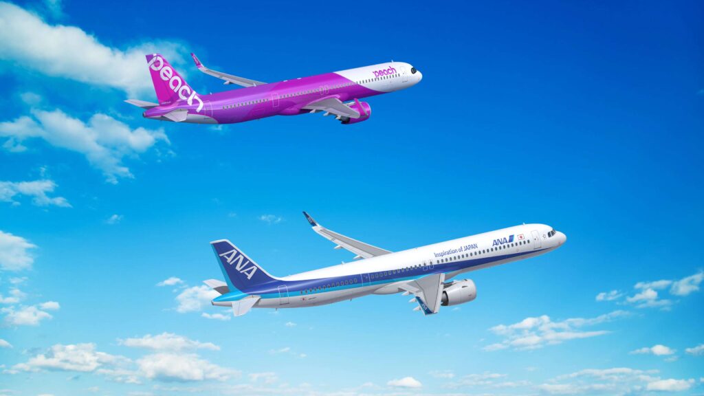 Two airplanes are flying in a clear blue sky with scattered clouds. The airplane on the top is painted in a bright pink and purple livery with the word "Peach" on its side. The airplane on the bottom has a white and blue livery with "ANA" and "Inspiration of JAPAN" written on it.
