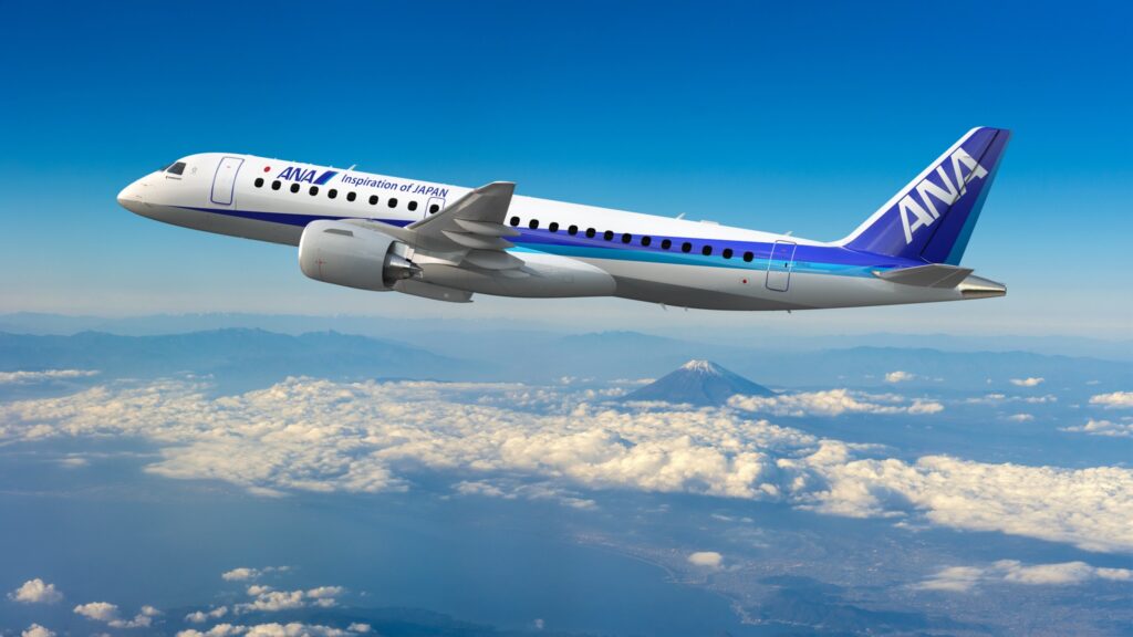 A passenger airplane with "ANA" and "Inspiration of JAPAN" written on its side is flying above the clouds. The aircraft is white with blue accents. In the background, there is a view of a mountain peak, possibly Mount Fuji, under a clear blue sky.