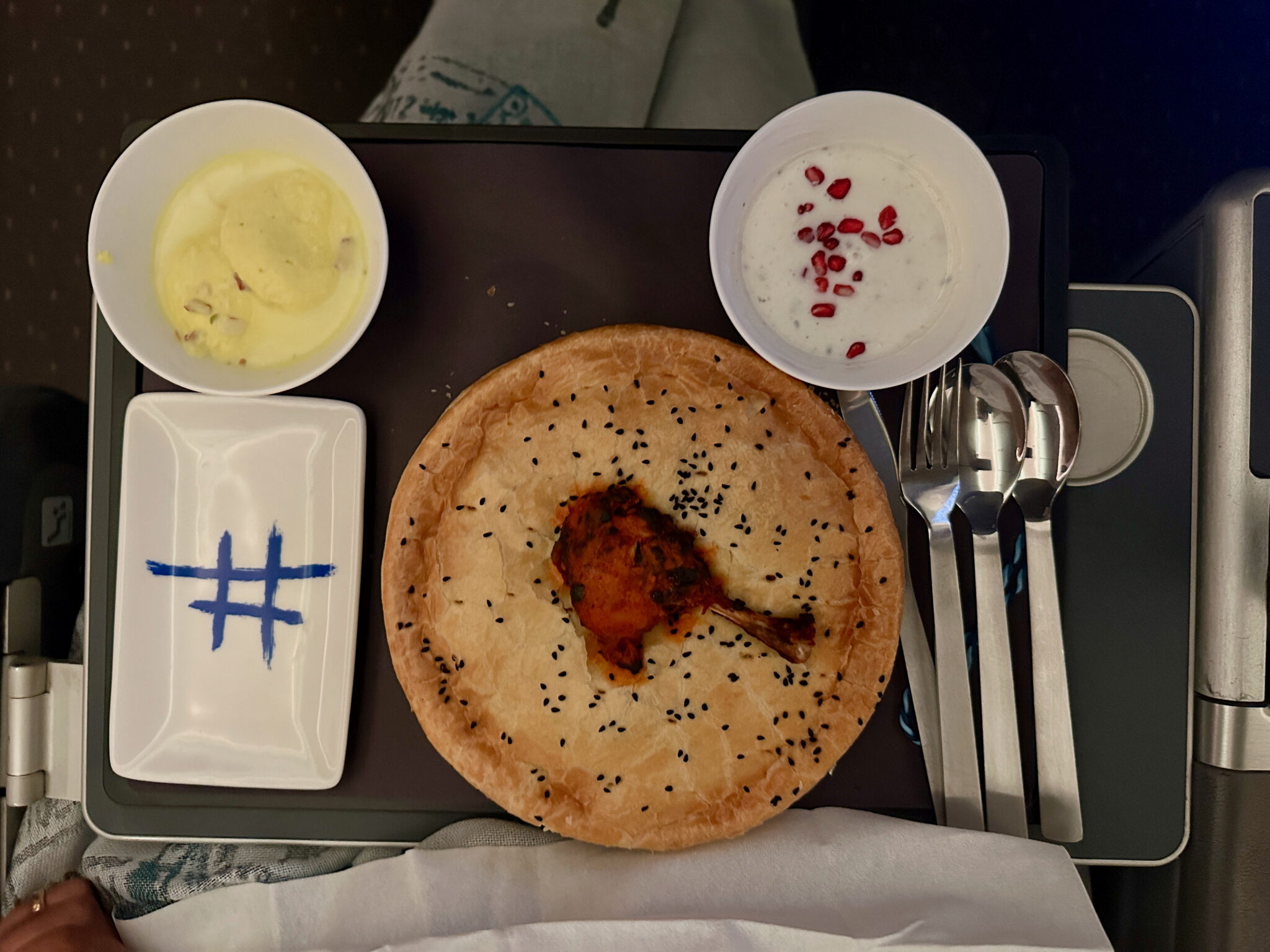 A meal is served on a tray, featuring a round pastry with black sesame seeds and a piece of chicken in the center. There are two small bowls: one with a creamy yellow dessert and the other with white yogurt topped with pomegranate seeds. A rectangular dish with a blue hashtag design is also present. Cutlery, including a fork, knife, and spoon, is placed on the right side of the tray.