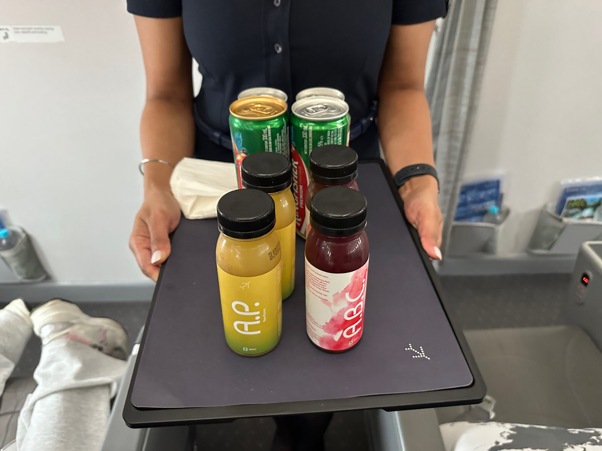 A person is holding a tray with various beverages on it, including bottles of juice and cans of soda. The person is wearing a dark uniform, and the setting appears to be inside an airplane, with seats and a passenger's leg visible in the background.
