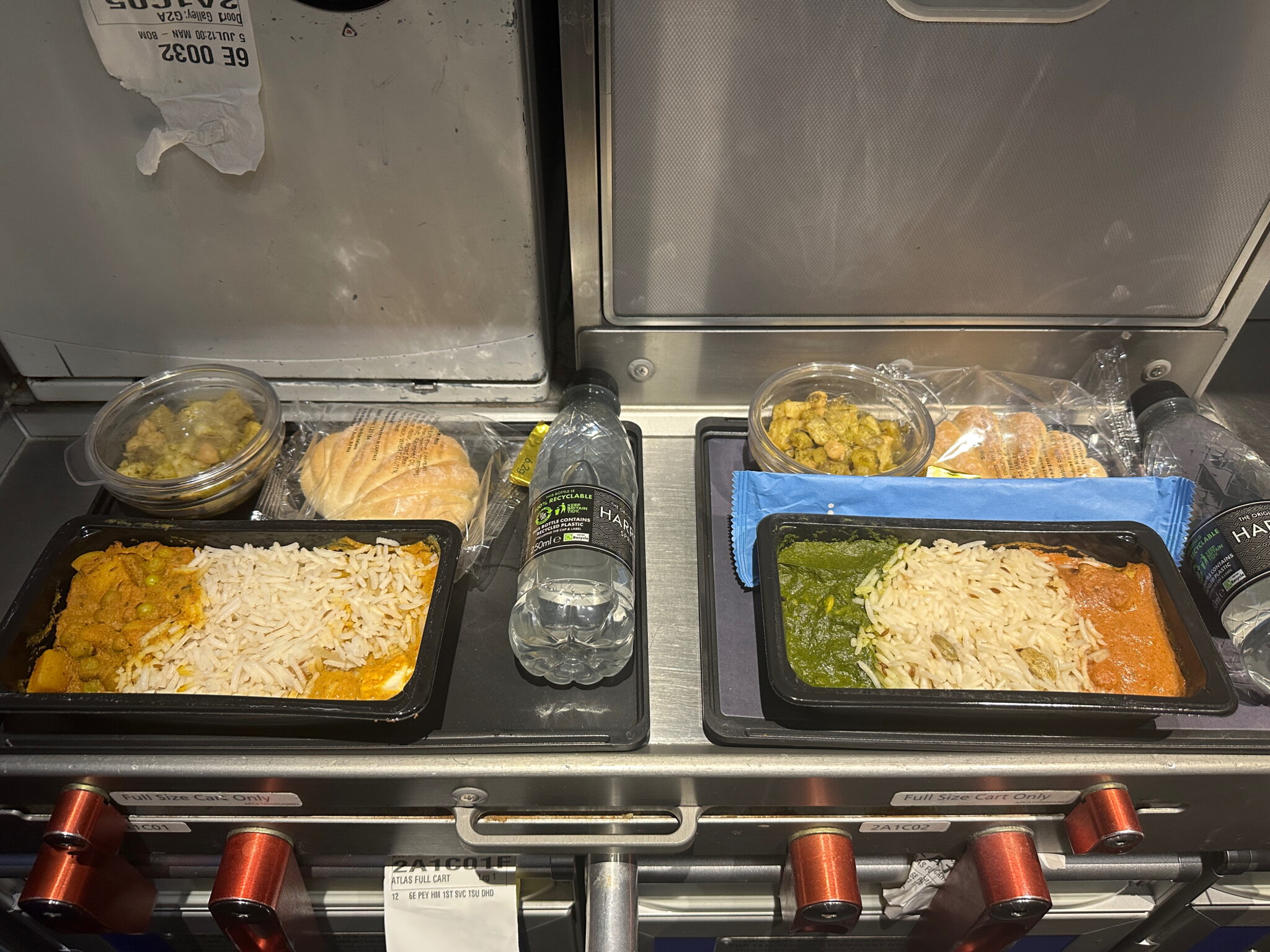 The image shows two trays of airplane meals placed on a metal surface. Each tray contains a compartmentalized meal with rice and curry dishes. There are also small bowls of what appears to be a vegetable side dish, packaged bread rolls, and bottles of water next to each tray. The setting suggests an in-flight meal service.
