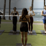 A group of people is practicing yoga on mats on a grassy area near the ocean. They are standing in a row, facing an instructor. Palm trees and waves are visible in the background. The text on the image reads, "Members Save 20%. Find last minute weekend getaways at over 2,800+ participating hotels and resorts." There are buttons labeled "Join Now" and "Terms Apply."