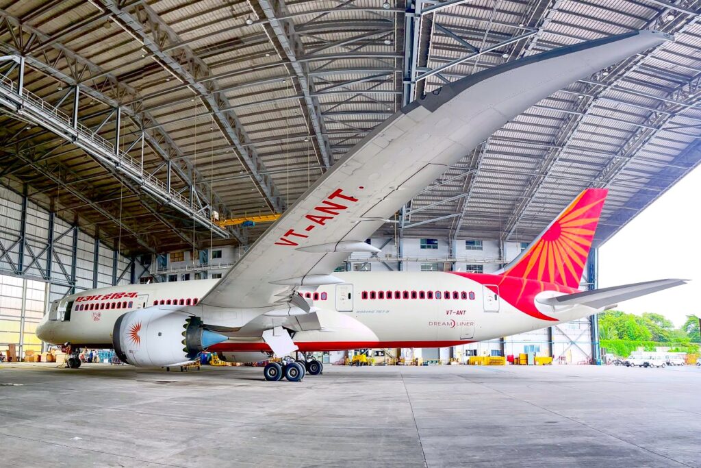 The image shows a large commercial airplane inside an aircraft hangar. The plane has a white body with red accents, including a red tail fin featuring a sunburst design. The registration number "VT-ANT" is visible on the wing. The hangar is spacious with a high ceiling and metal framework.