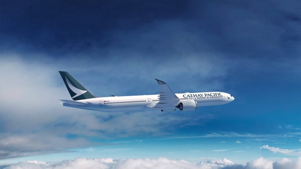 A Cathay Pacific airplane is flying through a clear blue sky above the clouds. The aircraft is white with the airline's logo and name visible on the side. The sky is bright with a few scattered clouds below.