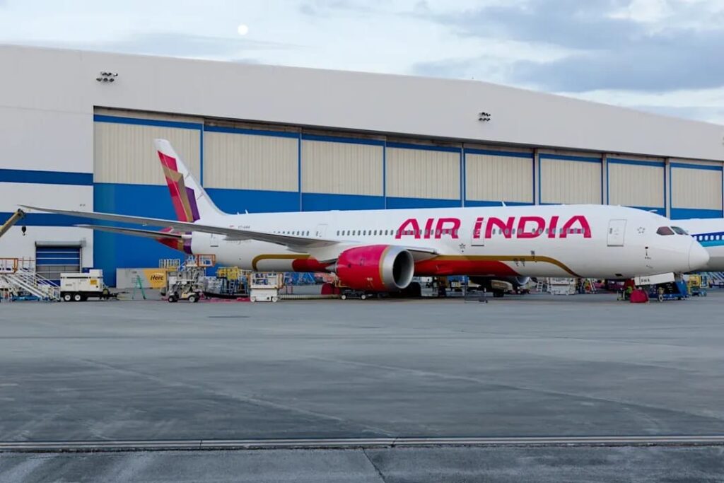 The image shows an Air India airplane parked on the tarmac in front of a large hangar. The aircraft is white with red and gold accents, and the airline's name is prominently displayed on the side. Various ground support equipment and vehicles are positioned around the plane. The sky is partly cloudy.