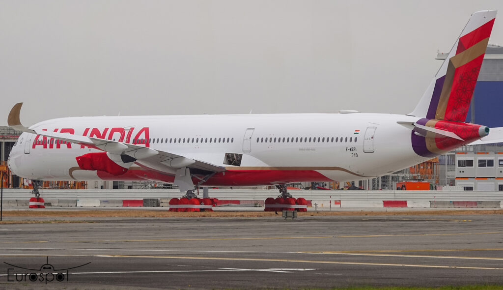 The image shows an Air India Airbus A350-1000 aircraft parked on the tarmac. The plane is white with red and gold accents, featuring the Air India logo on the fuselage. The tail has a colorful design with red, purple, and gold patterns. The aircraft is stationary, with its landing gear covered in red protective covers. The background includes airport buildings and infrastructure.