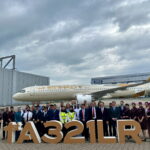 A group of people, including airline staff and executives, are standing in front of an Etihad Airways Airbus A321LR aircraft. They are posing for a photo with large letters in front of them spelling "1st A321LR." The background shows an Airbus hangar and a cloudy sky.