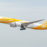 A yellow and white airplane with "flyscoot.com" written on the side is taking off. The aircraft has "Scoot" written on the tail and is in mid-air against a light gray sky.