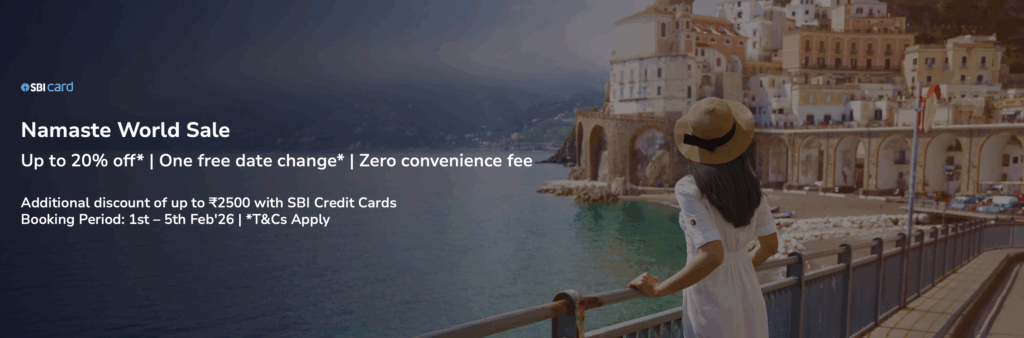 The image is an advertisement for the "Namaste World Sale" by SBI Card. It features a woman in a white dress and straw hat looking at a scenic coastal town with colorful buildings and arches. The text on the left side of the image offers up to 20% off, one free date change, and zero convenience fee. Additional discounts are available with SBI Credit Cards, and the booking period is from 1st to 5th February 2026. Terms and conditions apply.