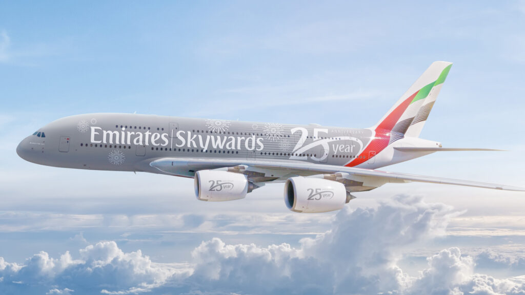 An Emirates Airbus A380 airplane is flying in the sky above the clouds. The aircraft is painted with a special livery celebrating the 25th anniversary of Emirates Skywards, featuring the text "Emirates Skywards 25 year anniversary" along the fuselage and engines. The tail fin displays the Emirates logo with red, green, and black colors.
