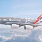 An Emirates Airbus A380 airplane is flying in the sky above the clouds. The aircraft is painted with a special livery celebrating the 25th anniversary of Emirates Skywards, featuring the text "Emirates Skywards 25 year anniversary" along the fuselage and engines. The tail fin displays the Emirates logo with red, green, and black colors.