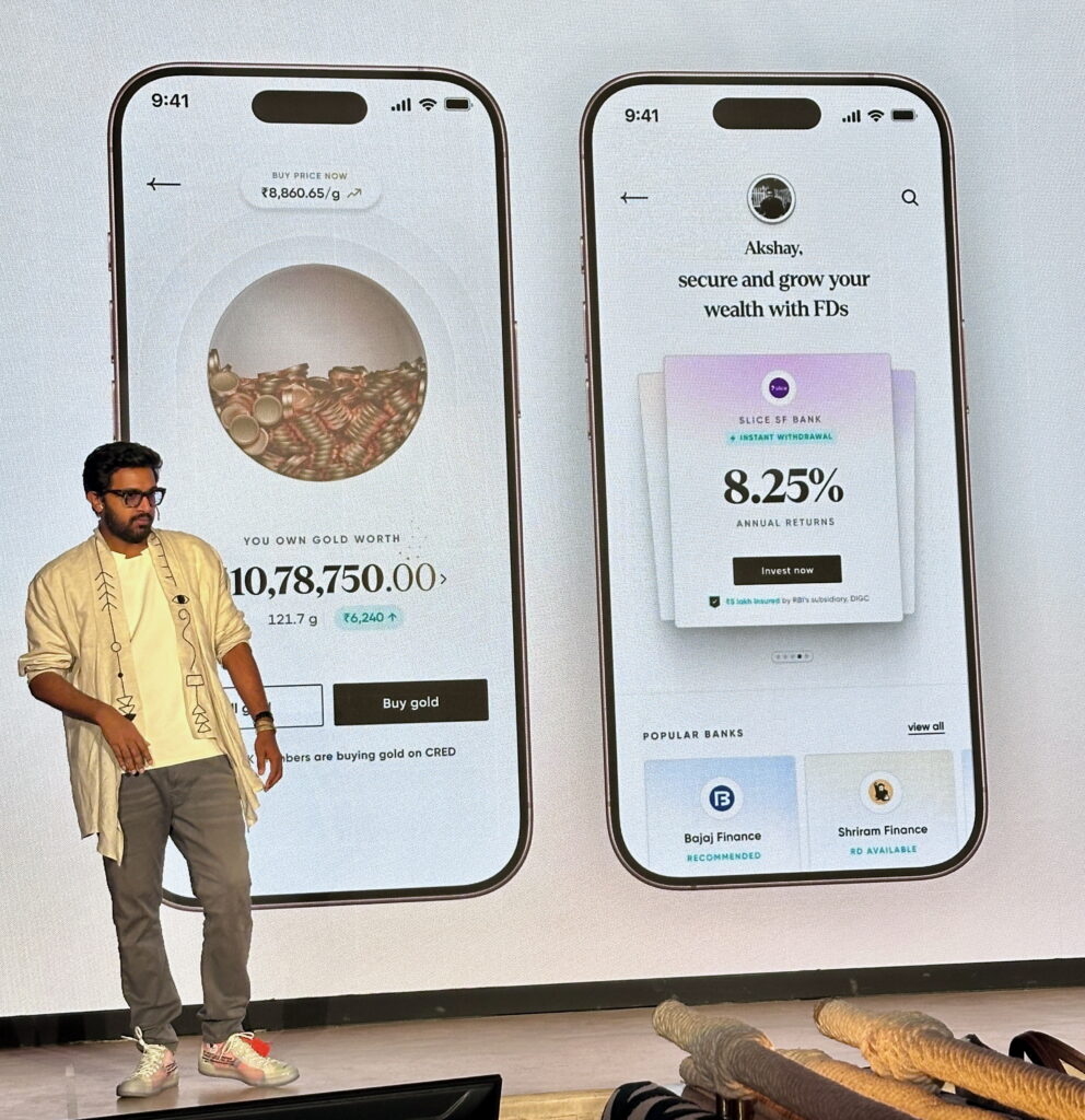 The image shows a person standing on a stage in front of a large screen displaying two smartphone screens. The left screen shows a gold investment app with details about gold ownership and current value. The right screen displays a financial app offering fixed deposit options with an 8.25% annual return. The person is wearing a light-colored jacket, white shirt, gray pants, and sneakers. There are ropes or cables visible at the bottom of the image.