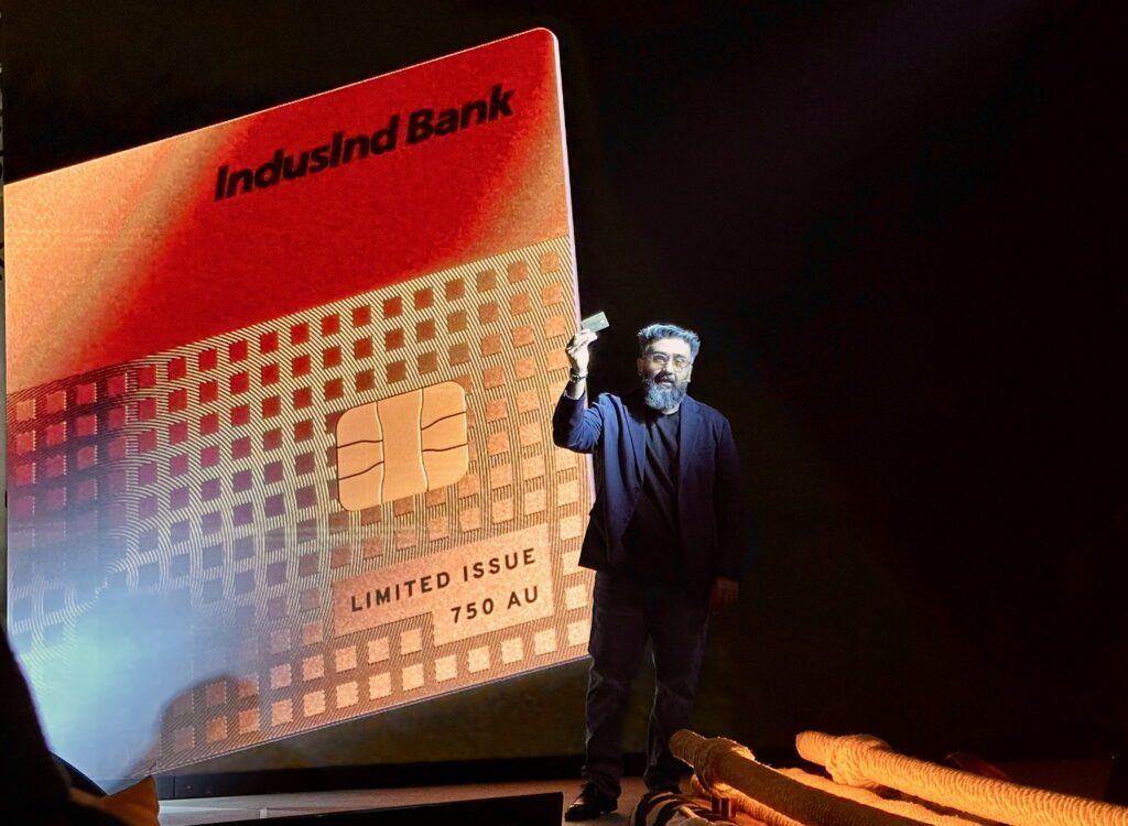 A man is standing on a stage holding a credit card. Behind him is a large projection of an IndusInd Bank card with a chip, labeled "LIMITED ISSUE 750 AU." The background is dark, and there are ropes visible on the stage.