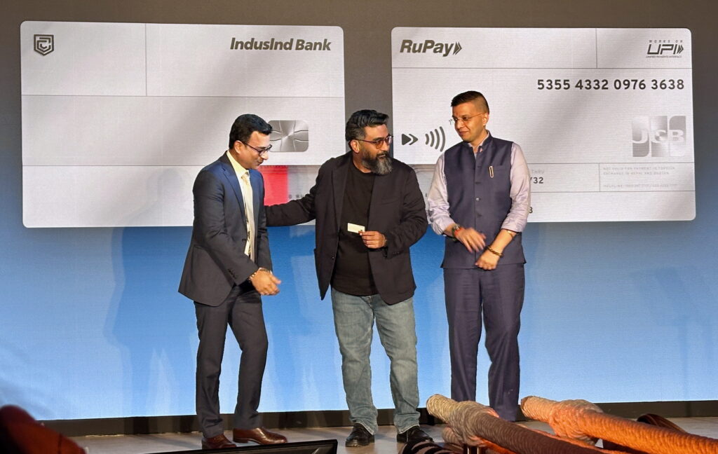 Three men are standing on a stage in front of a large backdrop featuring an image of a credit card. The card displays logos for IndusInd Bank, RuPay, UPI, and JCB. The man in the center is holding a small card, and all three are dressed in formal attire. The setting appears to be a formal event or presentation.