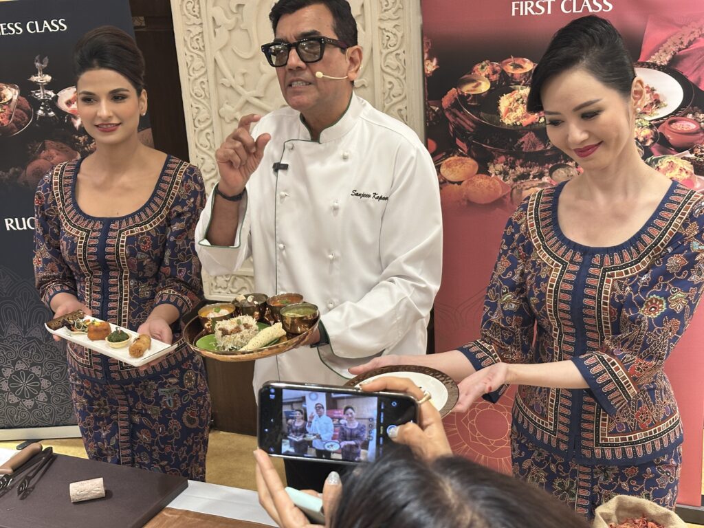 Three people are standing together in a promotional setting. The person in the center is wearing a chef's coat and holding a platter of food. The two people on either side are wearing matching patterned outfits and holding plates of food. A person in the foreground is taking a photo with a smartphone. The background features banners with images of food and text related to different classes of service.