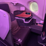 The image shows a luxurious airplane business class seat with a modern design. The seat is spacious and features a large, comfortable cushion with a patterned pillow. There is a personal entertainment screen, a side table with a control panel, and a window with a shade. The lighting in the cabin has a soft, purple hue, creating a relaxing atmosphere. A glass with a drink is placed on a small tray beside the seat.