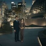 A man and a woman are standing together outdoors at night, dressed in formal attire. They are in an urban setting with modern architecture and illuminated skyscrapers in the background. The area is landscaped with trees and bushes, and the scene is well-lit, creating a sophisticated atmosphere.