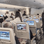 The image shows the interior of an airplane cabin with passengers standing and sitting. The seats are equipped with personal entertainment screens displaying various content. Some passengers are interacting with each other, and a few are holding drinks. The cabin is well-lit, and the overhead compartments are closed.