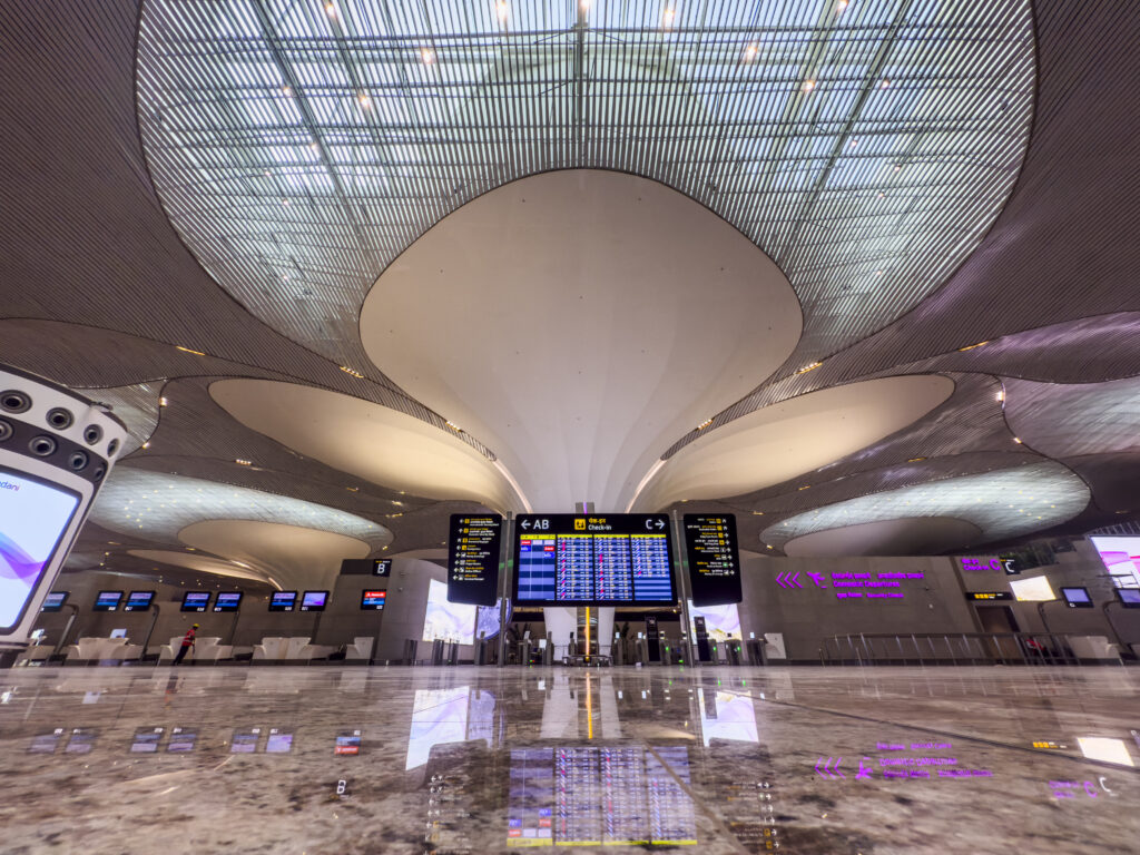 The image shows the interior of a modern airport terminal. The ceiling features a unique, wavy design with large, circular light fixtures. There are multiple digital screens displaying flight information and directions. The floor is shiny and reflective, and there are check-in counters visible in the background. The overall atmosphere is spacious and contemporary.