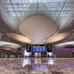 The image shows the interior of a modern airport terminal. The ceiling features a unique, wavy design with large, circular light fixtures. There are multiple digital screens displaying flight information and directions. The floor is shiny and reflective, and there are check-in counters visible in the background. The overall atmosphere is spacious and contemporary.