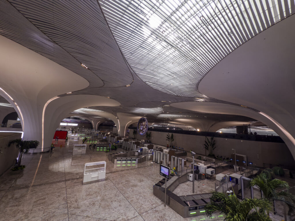 The image shows the interior of a modern airport terminal. The architecture features a large, open space with a high, curved ceiling and a pattern of lines that allow natural light to filter through. Below, there are security checkpoints with conveyor belts and scanning equipment. The floor is made of polished stone, and there are a few plants placed around the area. The overall design is sleek and futuristic.