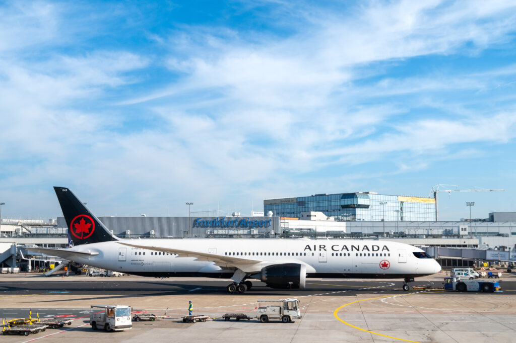 An Air Canada airplane is on the tarmac at an airport, with the terminal building in the background. The sky is clear with some clouds. Ground service vehicles and equipment are visible near the aircraft.