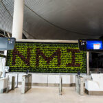 The image shows an airport gate area with the number 14 displayed on digital screens. In the background, there is a vertical garden with green and red plants arranged to form the letters "NMI." The area has modern design elements, including white counters and turnstiles. The ceiling has a curved, ribbed design.