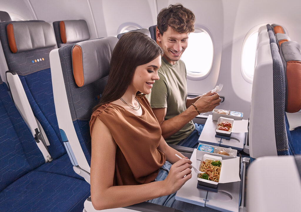 A man and a woman are sitting in airplane seats, enjoying in-flight meals. The woman is holding a fork and eating noodles, while the man is smiling and holding a fork as well. The airplane cabin is visible, with blue and gray seats and tray tables extended.
