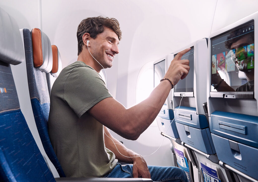 A man is sitting in an airplane seat, wearing earphones and smiling. He is interacting with the touchscreen of the in-flight entertainment system in front of him. The cabin interior is visible, including other seats and windows.