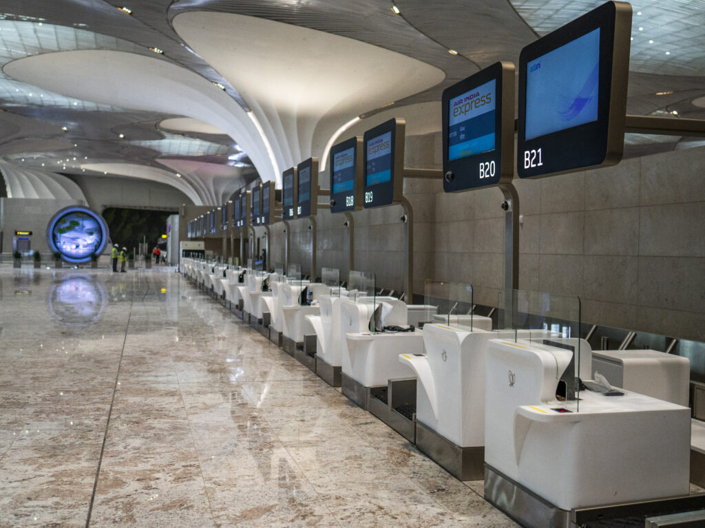 The image shows a modern airport check-in area with a row of empty check-in counters. Each counter has a digital screen displaying "Air India Express" and counter numbers like B19, B20, and B21. The area is spacious with a high ceiling and a polished marble floor. In the background, there are a few people and a large circular display. The overall atmosphere is clean and organized.