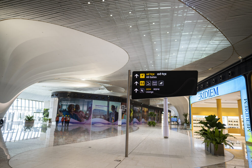The image shows a modern airport terminal with a sleek, curved ceiling design. In the foreground, there is a signpost with directions in both English and Hindi, indicating directions to "All Gates," "Multi Faith," and "Lounge." The terminal has a spacious, glossy floor and large windows on the left side. There are a few people walking in the distance, and a store with the name "MERIDIEM" is visible on the right. The area is well-lit and features some potted plants.