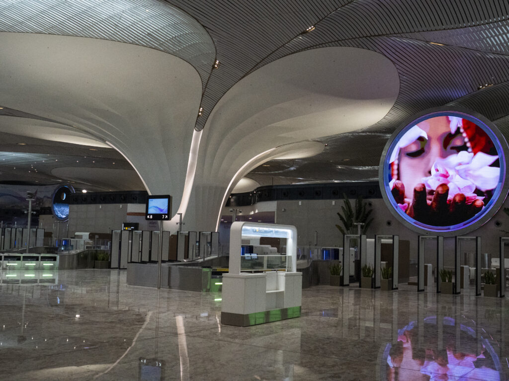 The image shows a modern airport interior with a spacious, sleek design. The ceiling features large, curved architectural elements. There is a digital display on the right side showing a close-up of a person holding flowers. The floor is shiny and reflective, and there are various counters and signs, including one indicating gates K8 and 7A. The area appears to be a check-in or security section with barriers and plants.