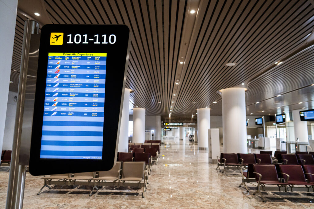 The image shows an airport terminal with a digital display board on the left. The board lists domestic departures, including flight numbers, destinations, gate numbers, and status, all marked as "On Time." The terminal has rows of empty seating and a high ceiling with lights. The floor is made of polished stone, and there are large white columns throughout the space.