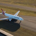 An airplane with "Qantas" written on the side is taxiing on a runway. The aircraft is white with red accents on the tail. The runway is surrounded by grassy areas, and the shadow of the plane is visible on the ground.