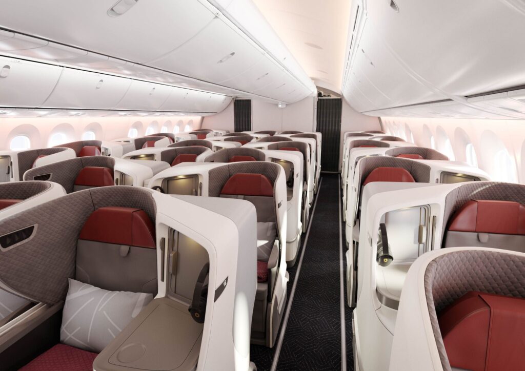The image shows the interior of an airplane's business class cabin. The seats are arranged in a staggered configuration, providing privacy and space for each passenger. The seats are upholstered in a combination of red and gray materials, with quilted patterns on the side panels. Each seat has a personal entertainment screen, a small table, and a pillow. The cabin is well-lit, with overhead compartments and windows along the sides. The aisle runs down the center, dividing the seating into two sections.