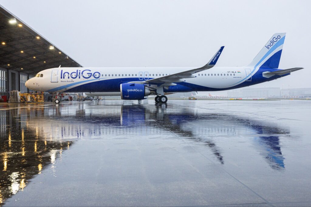 A large commercial airplane with "IndiGo" branding is parked on a wet tarmac near an airport hangar. The aircraft is painted in blue and white colors, and its reflection is visible on the wet surface. The sky is overcast, suggesting a rainy or cloudy day.