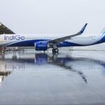 A large commercial airplane with "IndiGo" branding is parked on a wet tarmac near an airport hangar. The aircraft is painted in blue and white colors, and its reflection is visible on the wet surface. The sky is overcast, suggesting a rainy or cloudy day.
