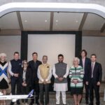 A group of thirteen people is standing in a line indoors, posing for a photo. They are dressed in formal and semi-formal attire. The setting includes two large white arches and a carpeted floor. In the foreground, there are two model airplanes on display.