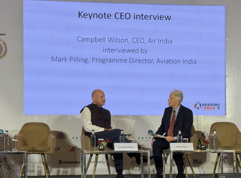 Two men are seated on a stage for a keynote CEO interview. The man on the left is wearing a vest and white shirt, and the man on the right is in a suit with a red tie. They are sitting in front of a large screen displaying text about the interview, which includes their names and titles. There are small tables with water bottles and flowers in front of them. The event is part of "Aviation India 2023."