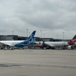 The image shows two airplanes parked at an airport terminal. The plane on the left has a blue and white color scheme with a distinctive logo on the tail. The plane on the right has a red and white color scheme with "Virgin" written on the tail. The airport tarmac is visible in the foreground, and the terminal building is in the background under a cloudy sky. Various ground service vehicles and equipment are positioned around the planes.