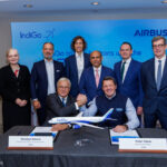 A group of eight people is posing for a photo in a conference room. Two men are seated at a table in the front, shaking hands over a model airplane. The table has nameplates for Christian Scherer and Pieter Elbers. Behind them, six people are standing, dressed in formal attire. A backdrop displays the logos of IndiGo and Airbus.
