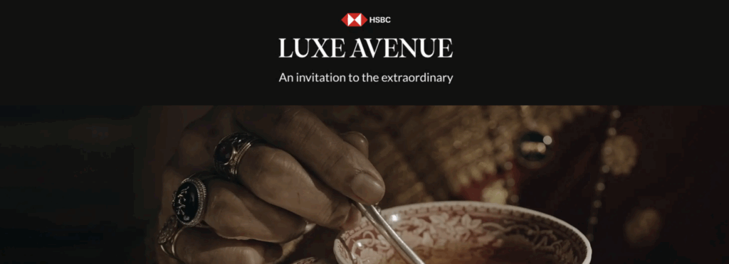 The image features a close-up of a hand holding a spoon over an ornate cup with a floral pattern. The hand is adorned with several rings. Above, there is a black banner with the HSBC logo and the text "LUXE AVENUE" followed by "An invitation to the extraordinary."